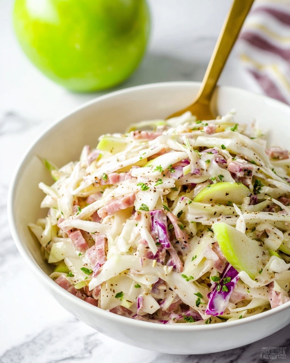 A white bowl filled with a creamy salad showing several layers including thin white cabbage strips, light pink pieces of ham, pale green apple slices, and small bits of purple cabbage mixed in. The salad has flecks of black pepper and chopped green herbs sprinkled on top. A gold spoon is stuck into the salad, standing upright at the back of the bowl. In the background, a green apple is partly visible on a white marbled surface. photo taken with an iphone --ar 4:5 --v 7
