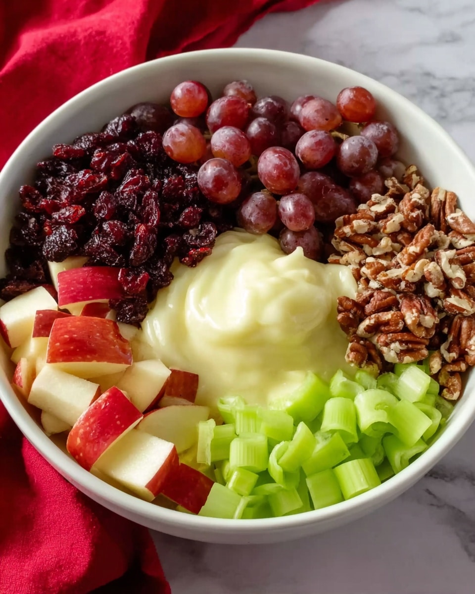 A white bowl is filled with six distinct layers arranged in a circle. Starting from the top left, there are small, dark red dried cranberries. Next to it, at the top center, are halved red grapes showing a light brown interior. To the right, finely chopped brown nuts are neatly piled. Below the nuts, bright green curved slices of celery rest in the bowl's lower right section. At the bottom left, small pieces of red apple with skin are visible. In the middle of the bowl sits a creamy, pale yellow dollop, smooth in texture and slightly glossy. The bowl is placed on a white marbled surface with a red fabric partially seen in the background. Photo taken with an iphone --ar 4:5 --v 7