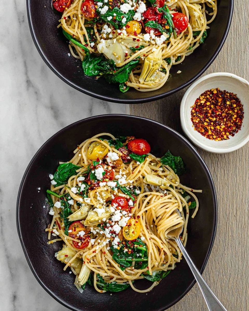 Two black bowls of spaghetti sit on a white marbled surface, each bowl filled with three main layers: a base of light golden spaghetti noodles, a middle layer of mixed vegetables including green spinach leaves, red and yellow cherry tomatoes, and pale artichoke pieces, and a top layer scattered with white crumbled cheese and red chili flakes. A silver fork rests in the bottom bowl with a small portion of pasta twirled around its tines. To the side, there is a small white bowl filled with crushed red pepper flakes. photo taken with an iphone --ar 4:5 --v 7