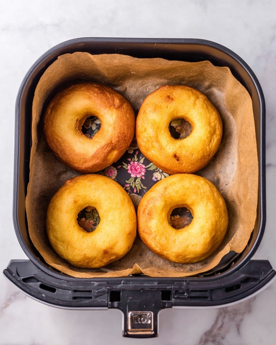 Four cooked golden-brown donuts with a smooth and slightly puffy texture are arranged in a square inside a black air fryer basket lined with brown parchment paper. The air fryer basket has a decorative floral pattern on the outside and is placed on a white marbled texture surface. The donuts have a consistent round shape with a hole in the center, showing slight variations in size and color, with some areas being lighter and others darker brown. Photo taken with an iphone --ar 4:5 --v 7