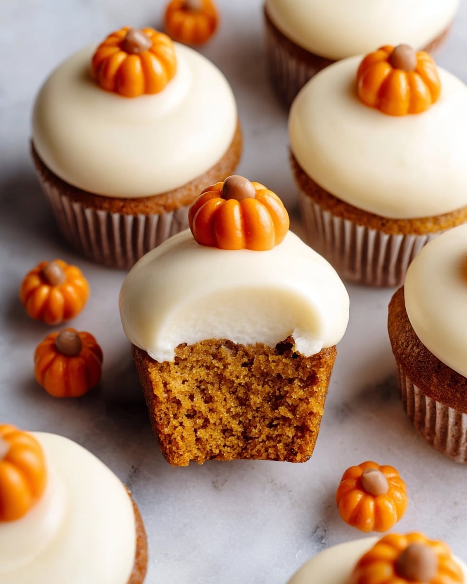 This image shows several small cupcakes arranged closely on a white marbled surface. Each cupcake has one layer of moist, orange-brown cake topped by a thick, creamy layer of smooth white frosting swirled on top in a round, puffed shape. On top of most cupcakes is a small, round orange pumpkin decoration with vertical grooves and a tiny brown stem in the center. One cupcake is cut in half, showing the soft crumb texture of the cake beneath the frosting. Scattered around the cupcakes are extra small orange pumpkin decorations. photo taken with an iphone --ar 4:5 --v 7
