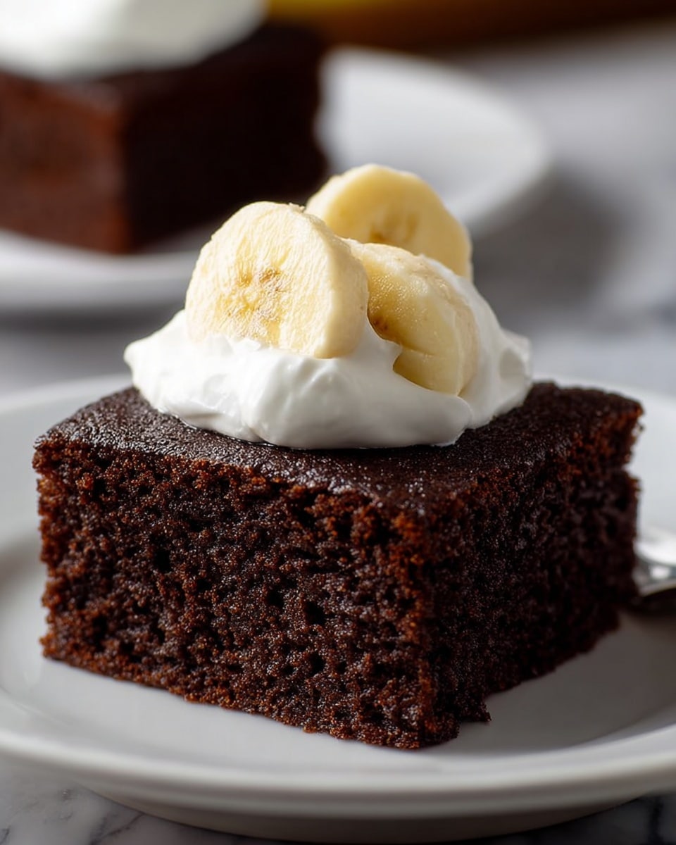 A thick square piece of dark brown chocolate cake with a moist and slightly crumbly texture sits centered on a white plate. On top of the cake is a dollop of smooth white whipped cream, with two thick slices of light yellow banana resting partly embedded in the cream. The background shows a blurred second plate with another piece of the same chocolate cake, all set on a white marbled surface. The photo is close up, highlighting the details of the cake's texture and the cream's softness. photo taken with an iphone --ar 4:5 --v 7