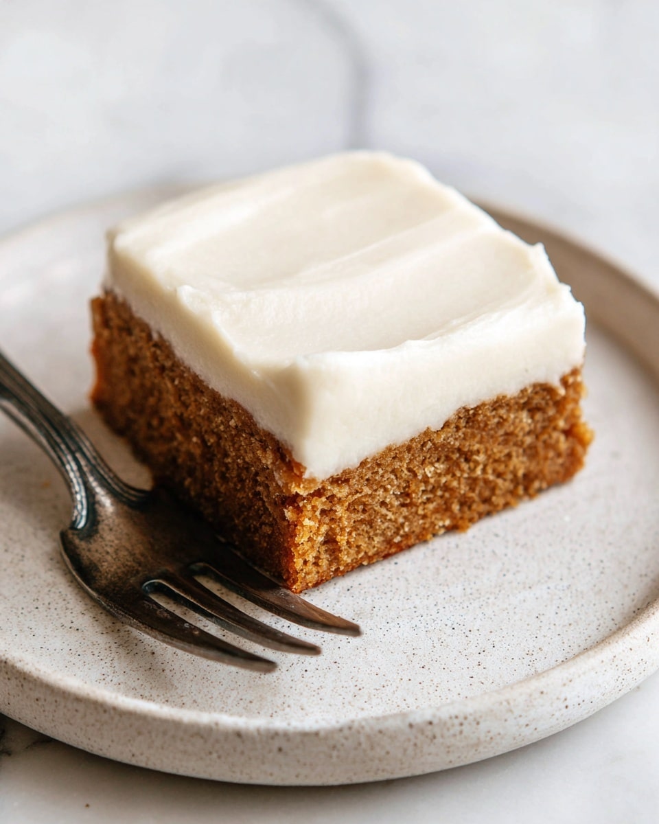 A single square piece of cake sits on a white plate with a slightly rough texture. The cake has two visible layers: a thick, dense, light brown bottom layer with a coarse texture, and a thick, smooth, creamy white frosting layer on top that covers the entire surface evenly with soft, slightly uneven edges. To the left of the cake, a silver fork rests partly on the plate. The scene is set on a white marbled surface. Photo taken with an iphone --ar 4:5 --v 7