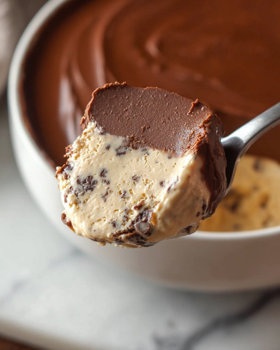 A close-up of a spoon holding a scoop of a two-layer dessert, with a thick, smooth dark brown chocolate layer on top and a light beige creamy layer with small dark specks underneath, showing a soft and slightly crumbly texture; in the background, a white bowl containing more of the beige creamy layer is visible on a white marbled surface. photo taken with an iphone --ar 4:5 --v 7