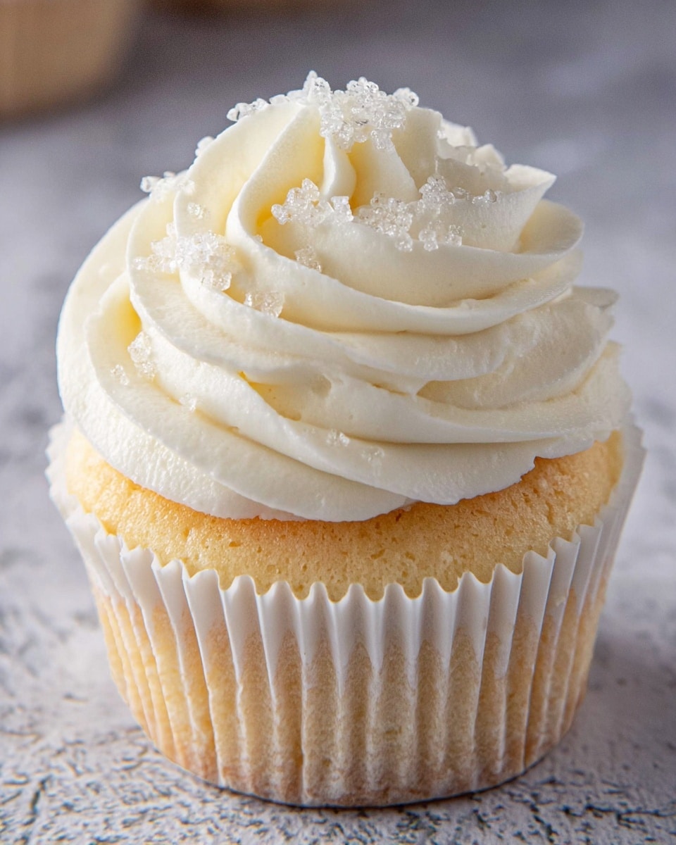A close-up of a single cupcake with two layers: the bottom layer is a light golden-yellow cake wrapped in a white fluted paper liner, and the top layer is a generous swirl of smooth, creamy white frosting decorated with small, clear sugar crystals, all resting on a white marbled texture surface. photo taken with an iphone --ar 4:5 --v 7