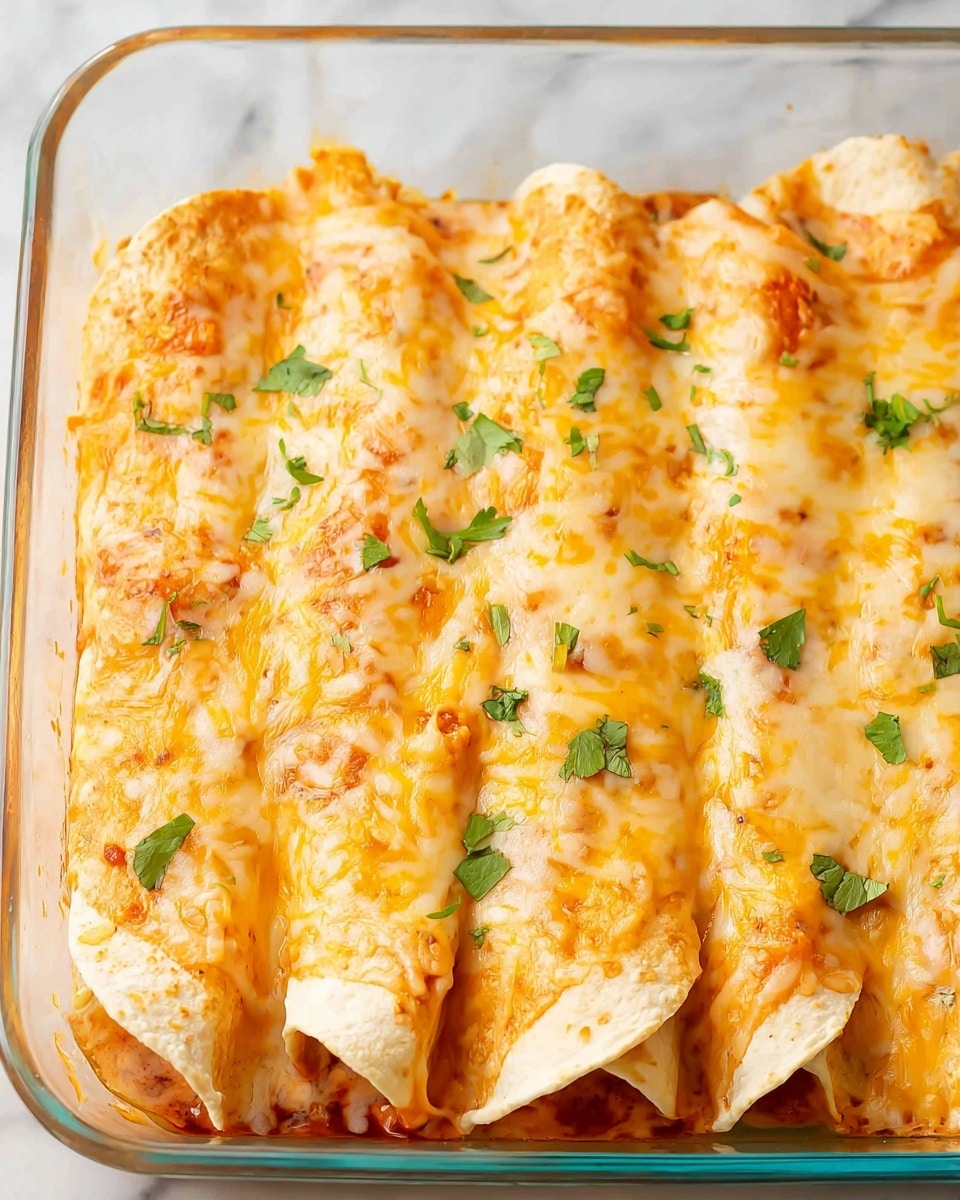 The dish shows five rolled enchiladas placed side by side inside a clear glass baking dish. Each enchilada is fully covered in melted, golden-yellow cheese with a slightly bubbly texture and has small green bits of fresh cilantro sprinkled on top. The wrapped tortillas beneath appear soft, slightly orange from sauce, and blend with the cheese layer. The clear baking dish reveals a red sauce layer underneath the enchiladas, visible along the edges. The setting is on a white marbled surface. photo taken with an iphone --ar 4:5 --v 7