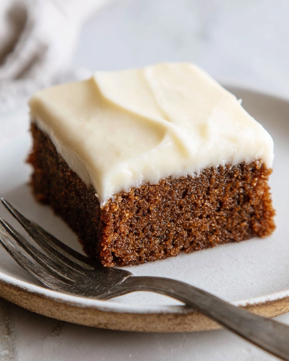 A single square piece of moist brown cake with a rough textured base layer topped with a thick, smooth, creamy white frosting layer, perfectly even and covering the entire top of the cake. The cake sits on a white plate with a slight natural edge texture, next to a metal fork on the left side. The background is a white marbled surface with soft natural light. Photo taken with an iphone --ar 4:5 --v 7