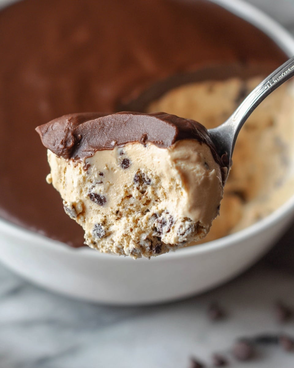 A close-up image of a spoon holding a creamy dessert scoop with two visible layers: the bottom layer is light beige with small dark bits inside, showing a smooth and soft texture; on top is a thick, shiny, and slightly cracked dark brown chocolate layer covering part of the beige layer. The background shows the remains of the dessert inside a clear bowl, resting on a white marbled surface. Photo taken with an iphone --ar 4:5 --v 7
