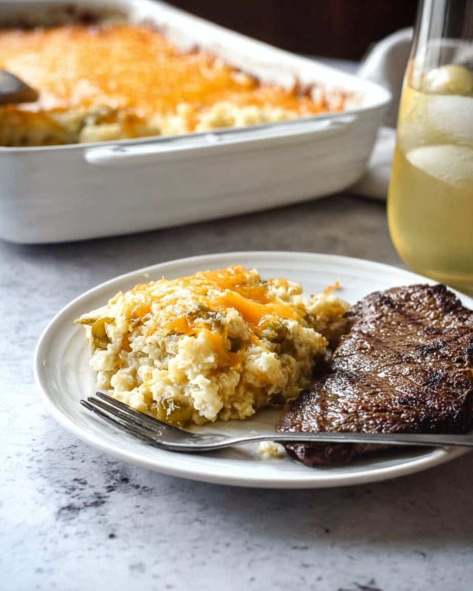 On a white plate, there is a serving of creamy casserole with three visible layers: a soft white rice or grain base, a middle layer with small pieces of light green vegetables, and a top layer melted golden and white cheese slightly browned. Next to the casserole is a thick, cooked steak with a dark brown charred texture. A metal fork rests on the left side of the plate, angled toward the casserole. In the background, there is a white casserole dish filled with more of the same casserole and a clear glass filled with light yellow beverage, all set on a white marbled texture surface with a beige burlap cloth underneath the plate. Photo taken with an iphone --ar 4:5 --v 7