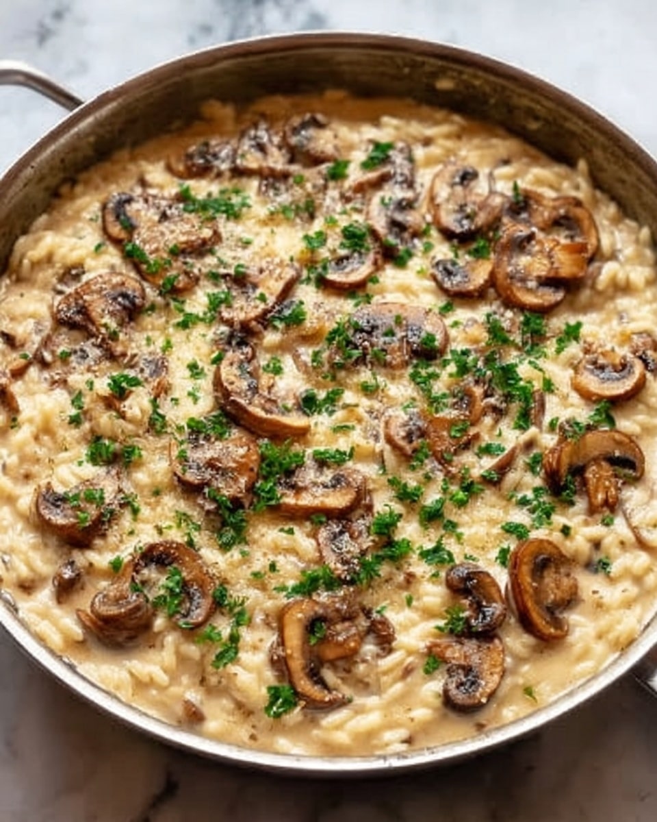 A close-up view of a creamy mushroom risotto in a shiny stainless steel pan. The dish shows a thick, creamy beige rice base mixed with slightly browned, soft slices of mushrooms all over. Small pieces of fresh green herbs are scattered on top, adding a touch of bright green color to the smooth, rich texture. The background is a white marbled surface, making the pan and food colors stand out clearly. Photo taken with an iphone --ar 4:5 --v 7