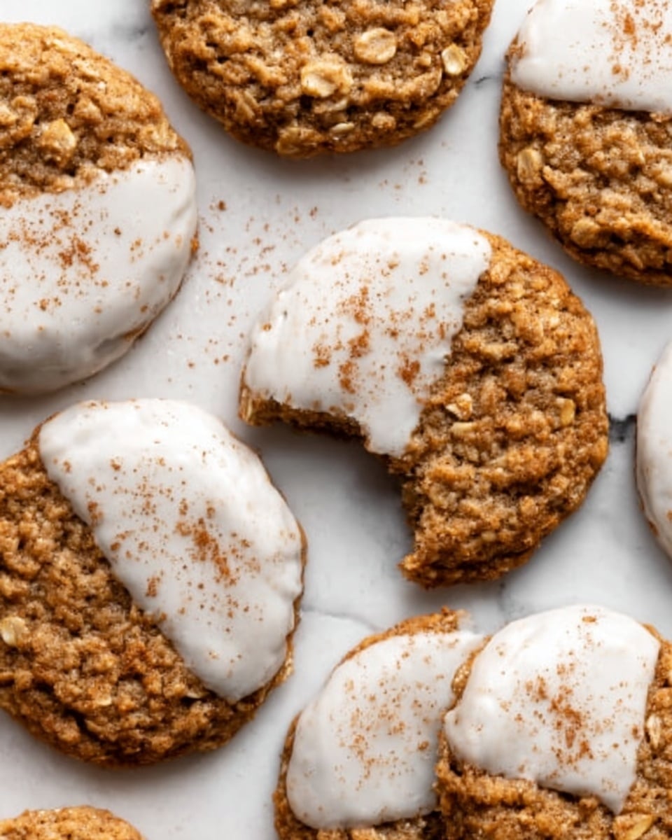 A group of round oatmeal cookies, some dipped halfway into white icing and sprinkled with a light dusting of cinnamon, are spread out on a white marbled surface. The cookies have a rough, textured surface with visible oats and a golden-brown color. One cookie shows a bite taken out of it, revealing a soft, chewy inside. The cookies are arranged casually, overlapping slightly, with the contrast of the creamy white icing and the warm brown cookie creating a cozy, inviting look. Photo taken with an iphone --ar 4:5 --v 7