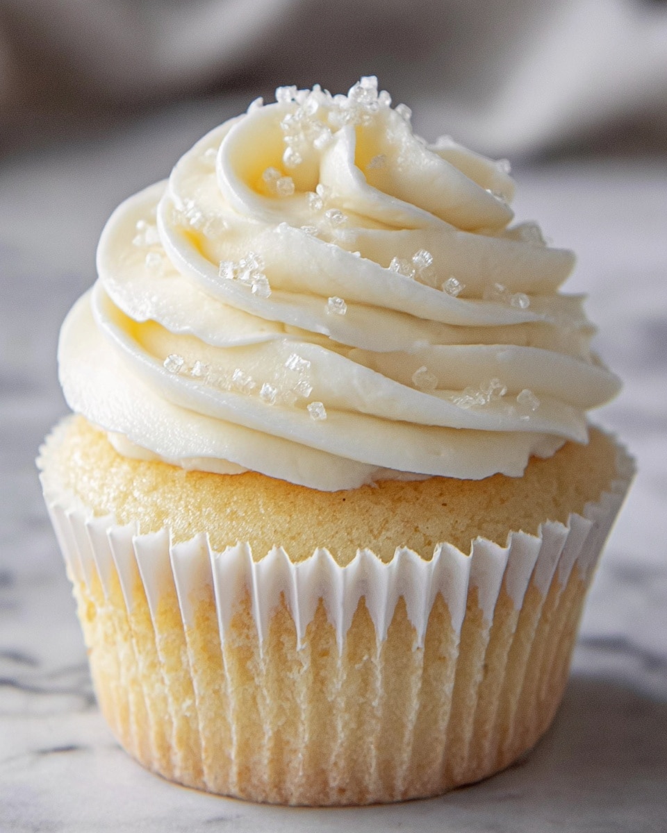 A single cupcake sits in a white pleated paper liner, showcasing a light golden cake base topped with a thick swirl of creamy white frosting. The frosting is textured with smooth, curving waves that build up three visible layers, decorated with small, clear sugar crystals that catch the light. The cupcake rests on a white marbled surface, creating a clean and simple backdrop that highlights the soft, delicate details of the cupcake. photo taken with an iphone --ar 4:5 --v 7