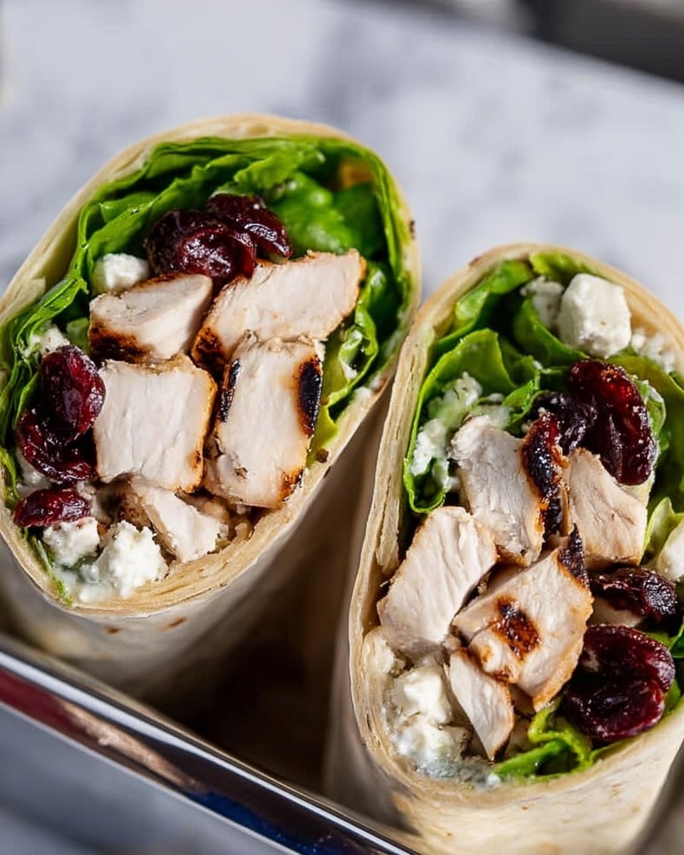 Two halves of a wrap are shown, each cut to display layers inside. The outer layer is a light beige tortilla wrap, tightly rolled. Just inside, there is a layer of green leafy lettuce, crisp and bright. The main filling consists of thick, juicy slices of white grilled chicken with a slight brown sear. Interspersed among the chicken are bits of white, crumbly cheese and dark red dried cranberries, adding texture and color contrast. The wrap halves rest in a shiny silver holder, and the background features a white marbled texture. photo taken with an iphone --ar 4:5 --v 7