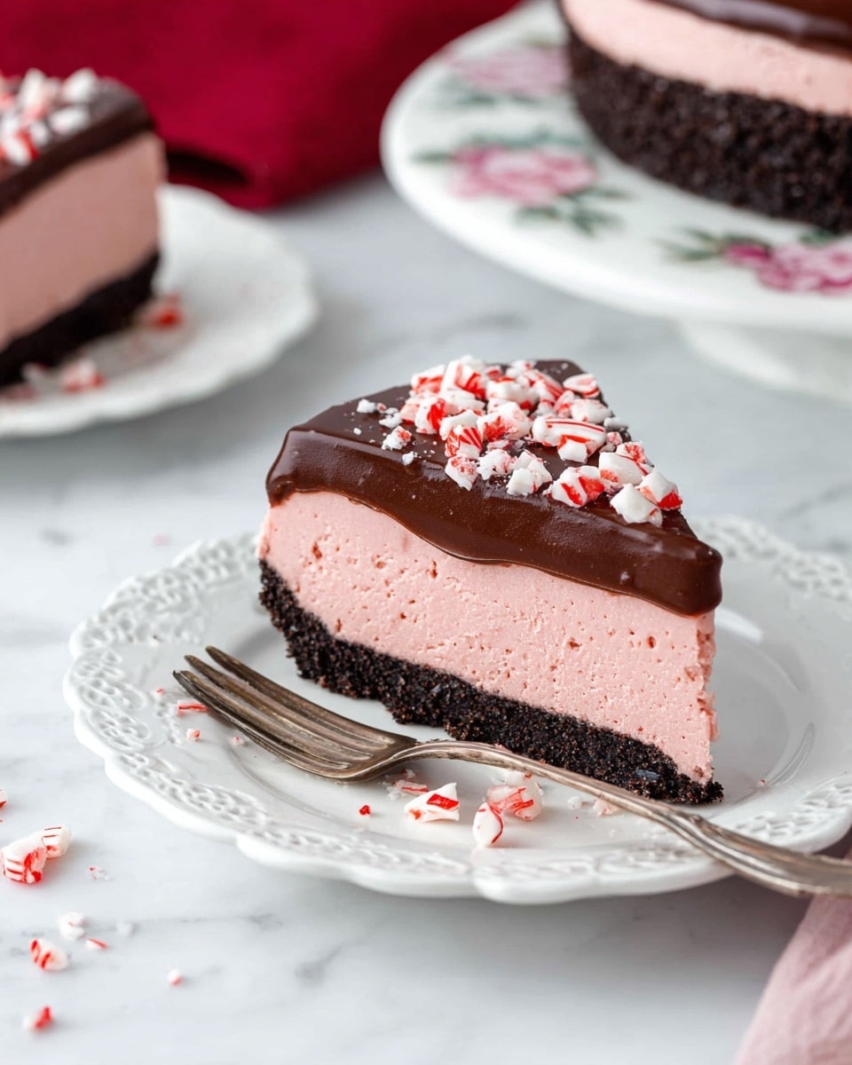 A slice of three-layer dessert is shown on a white plate with delicate lace-like edges. The bottom layer is a dark chocolate crumb crust, firm and textured. Above it, there is a thick, smooth, light pink mousse layer that looks creamy and soft. On top, there is a glossy dark chocolate ganache that covers the mousse, sprinkled with broken pieces of red and white peppermint candy. Next to the plate, a vintage silver fork lies on the white marbled surface, along with some scattered peppermint pieces. In the background, part of the whole dessert is visible on a white plate with a floral pattern, and a soft red cloth adds a touch of color. Photo taken with an iphone --ar 4:5 --v 7
