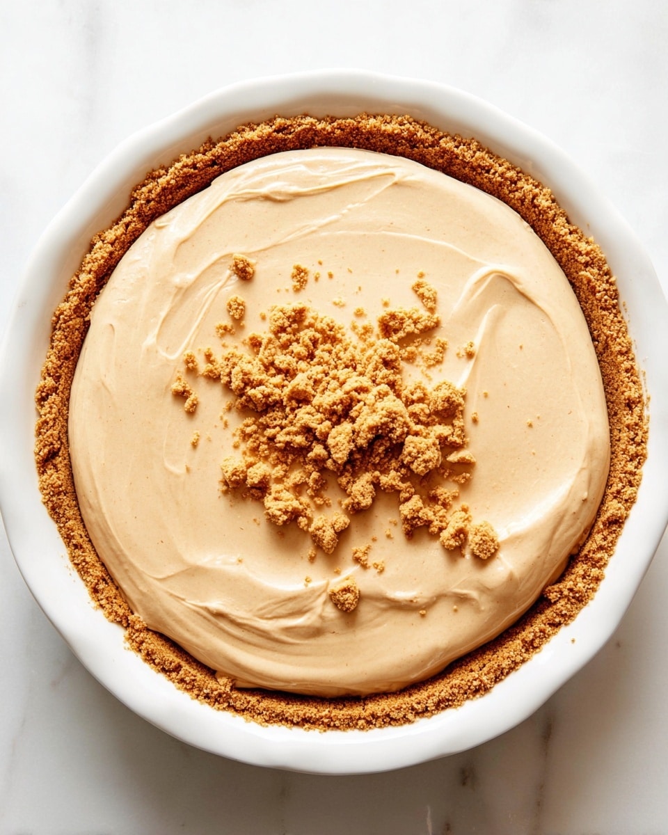 A round dessert with two visible layers: the bottom layer is a thick, crumbly, golden brown crust that lines the base and edges of a white pie plate, and the top layer is a smooth, creamy light beige filling spread evenly across the crust. In the center, there are small clusters of crushed crumbs scattered on the creamy surface, adding texture contrast. The whole pie sits on a white marbled surface. photo taken with an iphone --ar 4:5 --v 7