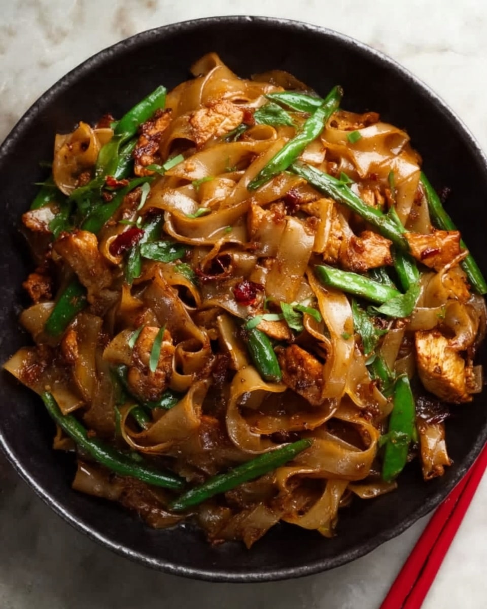 A black pan filled with wide, flat noodles mixed with cooked pieces of chicken and green vegetables like slices of green onion and green beans, all coated in a shiny, dark brown sauce. The noodles have a soft texture with a few curled edges, sitting on a white marbled surface with a few red chopsticks partly visible on the right side. The dish looks hot and fresh with some bits of herbs sprinkled on top. Photo taken with an iphone --ar 4:5 --v 7
