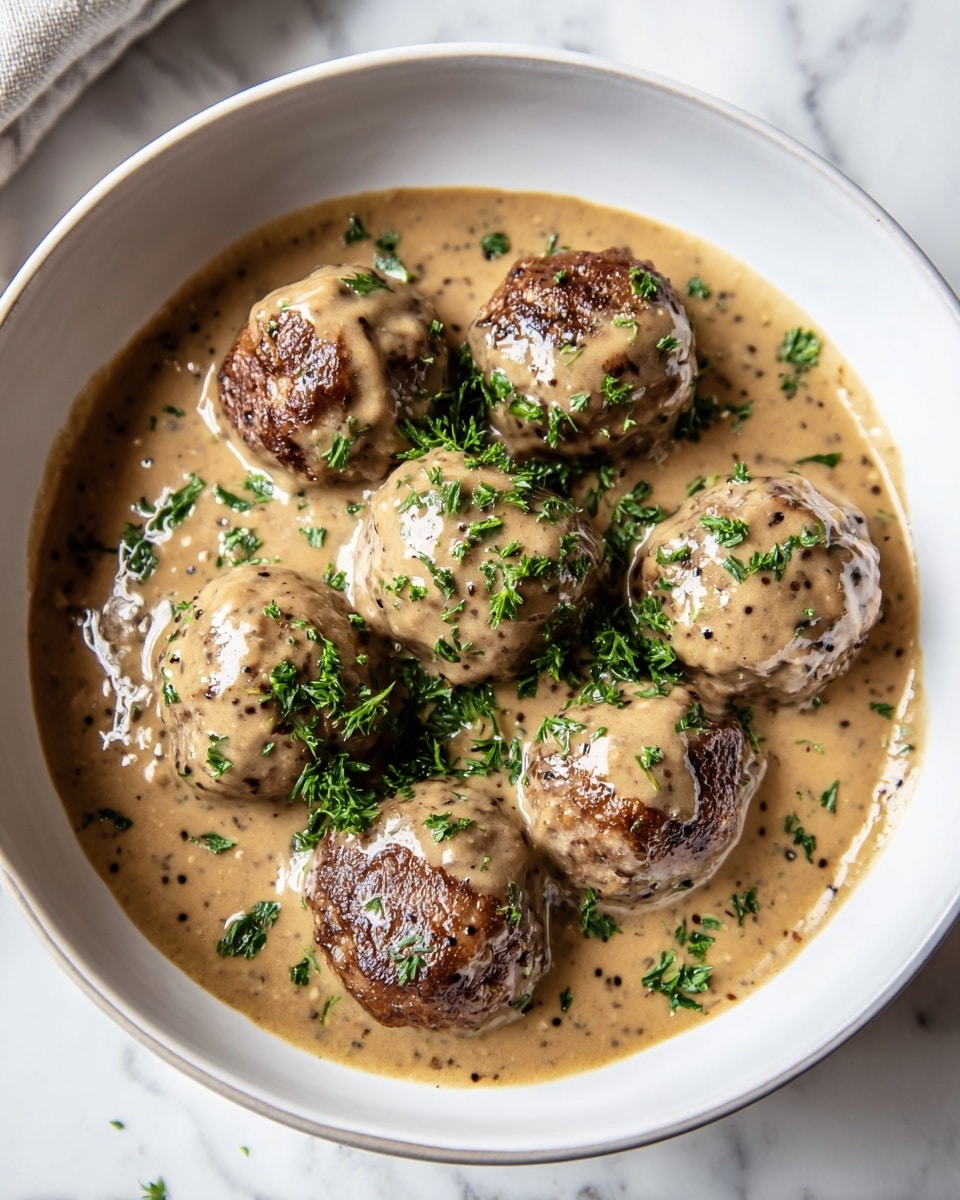A white bowl filled with seven brown meatballs covered in a thick, creamy light brown sauce, scattered with bright green chopped herbs on top and around the meatballs. The meatballs have a slightly rough texture with some visible browned spots, sitting in a smooth sauce that pools at the bottom of the bowl. The bowl rests on a white marbled surface. photo taken with an iphone --ar 4:5 --v 7