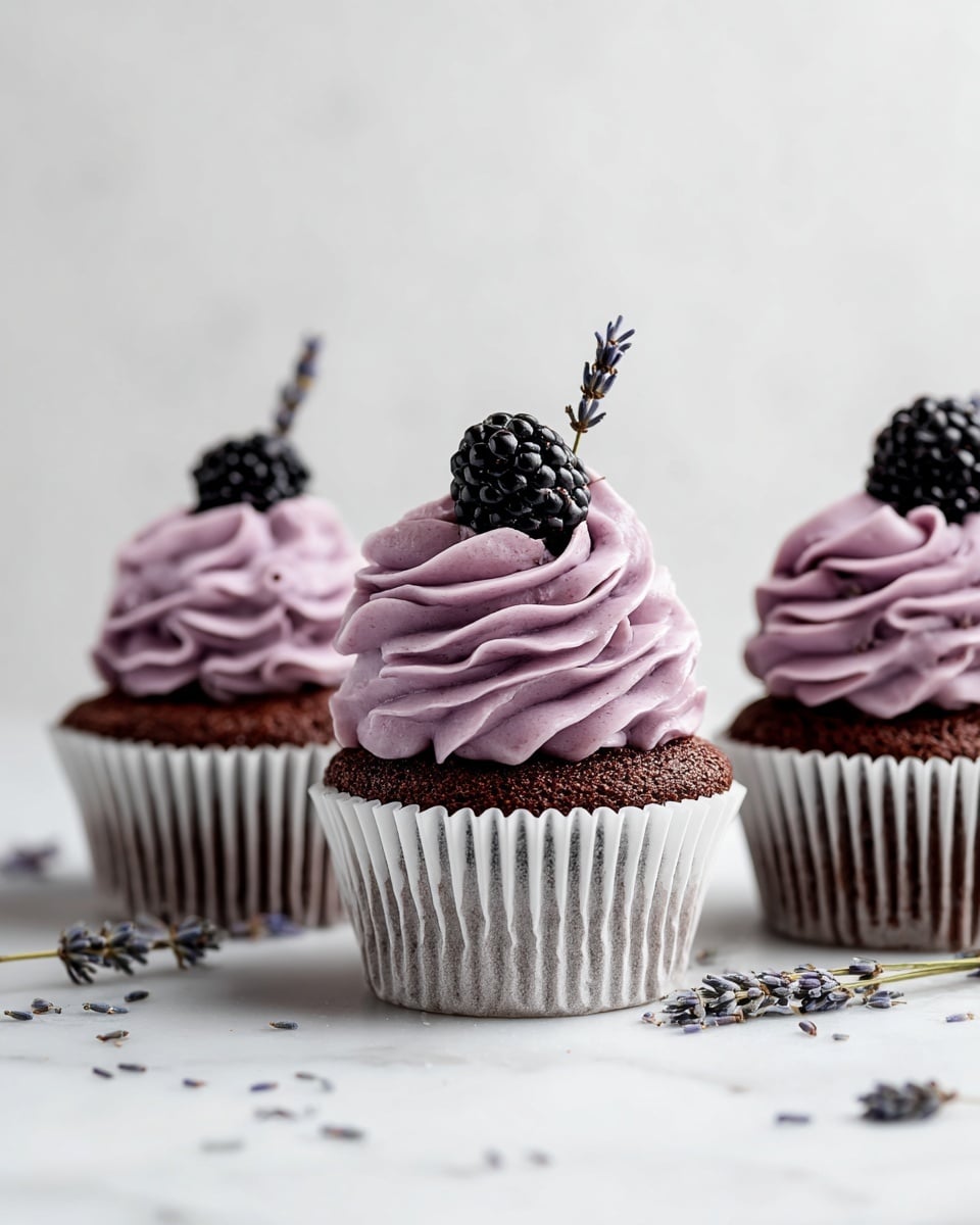 Three chocolate cupcakes sit side by side on a white marbled surface, each wrapped in brown paper liners. Each cupcake has a thick, swirled layer of light purple-pink frosting with a slightly textured appearance, topped with a shiny black blackberry and a small sprig of dried lavender. The background is plain and light, letting the cupcakes stand out clearly. Photo taken with an iphone --ar 4:5 --v 7