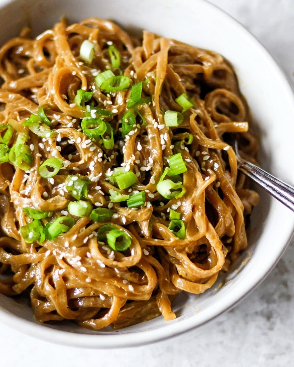 A close-up of a white bowl filled with thick flat noodles coated in a creamy brown sauce, topped with chopped bright green onions and sprinkled with white sesame seeds scattered evenly across the noodles. A silver fork rests inside the bowl on the right side, with noodles twirled around it, all placed on a white marbled surface. The noodles look soft and glossy with a slightly rough texture from the sauce. photo taken with an iphone --ar 4:5 --v 7