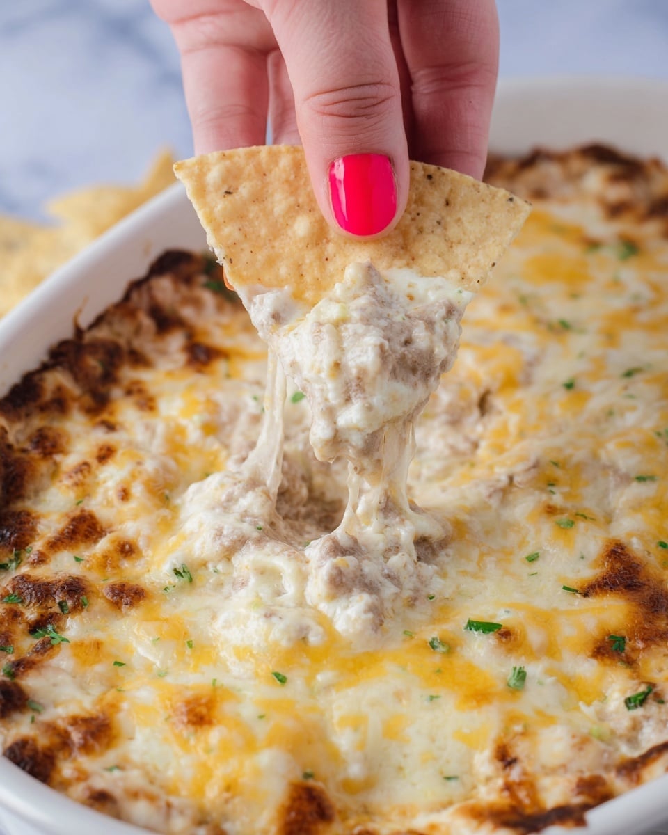 A close-up view of a white rectangular dish filled with a creamy, melted cheese dip mixed with beans, showing a textured surface with patches of golden brown melted cheese. A woman's hand with bright red painted nails holds a triangular tortilla chip dipped in the creamy mix, lifting it from the dish. The dip has a slightly chunky texture with visible bits of beans and cheese melted unevenly across the top. The background has a white marbled texture and a hint of blue is visible at the edge. photo taken with an iphone --ar 4:5 --v 7