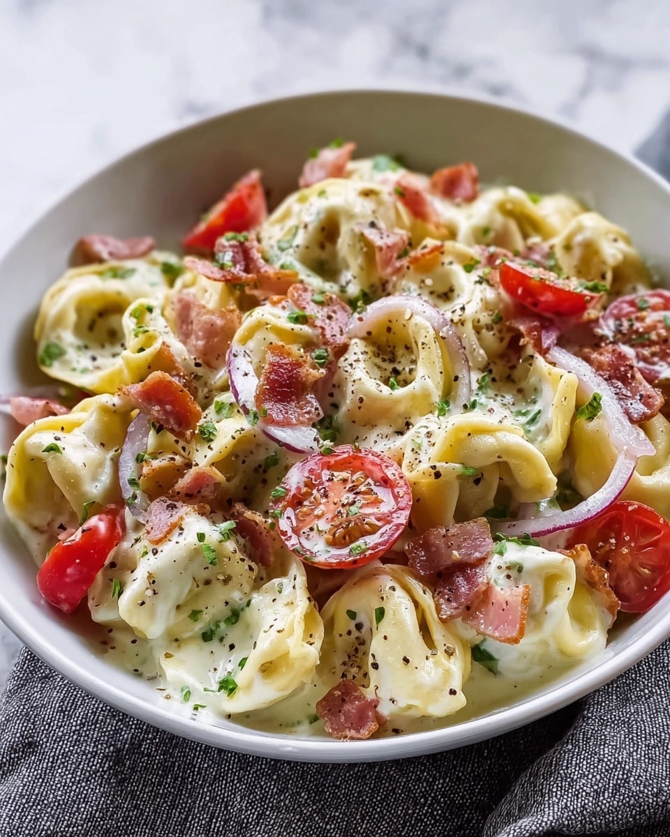 A close-up view of a white bowl filled with creamy tortellini pasta. The tortellini are pale yellow with a smooth texture, covered in a white sauce. Mixed within the pasta are bright red halved cherry tomatoes, thin slices of light purple onion, and small pieces of crispy brown bacon. The dish is sprinkled with finely chopped green herbs and black pepper flakes, adding texture and color. The bowl is placed on a soft gray cloth over a white marbled surface. Photo taken with an iphone --ar 4:5 --v 7