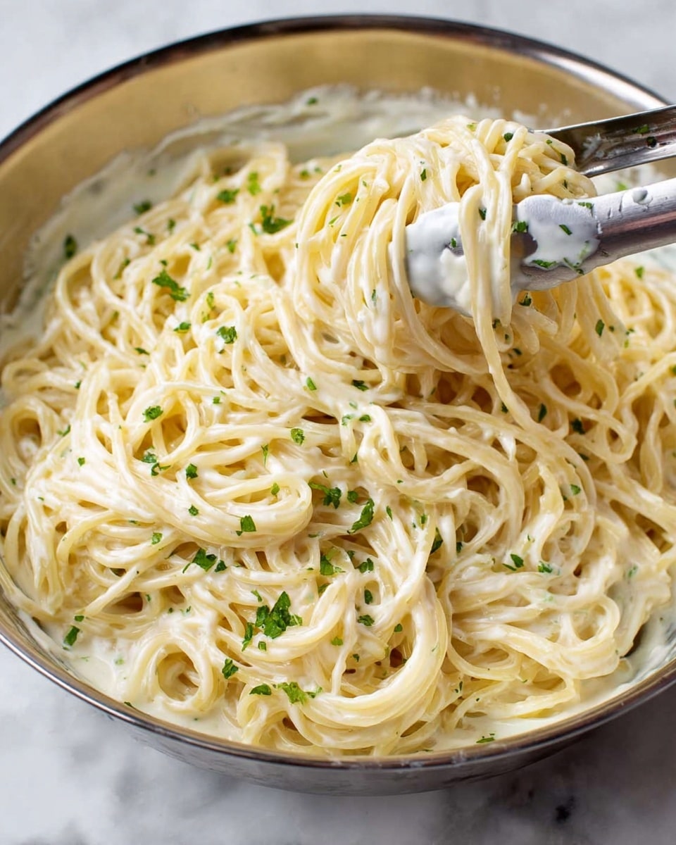 A close-up view of creamy spaghetti pasta coated in a smooth, light yellow sauce, with small green parsley pieces scattered on top for color contrast. The pasta strands are long, thin, and tangled together, held by silver tongs lifted from a stainless steel bowl that shows traces of the sauce on the sides. The background is a white marbled texture. photo taken with an iphone --ar 4:5 --v 7