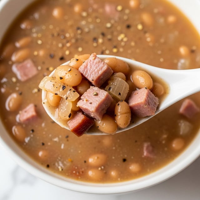 A close-up image showing a white spoon lifting a scoop of white beans mixed with diced ham and soft cooked onions in a thick, light brown broth. Black pepper is sprinkled on top of the beans and broth, adding small dark specks. The beans are creamy and oval-shaped, while the ham pieces are pink with a slightly firm texture. The background is a white marbled surface that softly blurs out, focusing attention on the spoonful of beans and meat. photo taken with an iphone --ar 4:5 --v 7