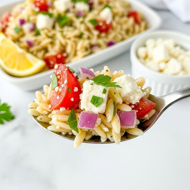 A close-up view of a silver spoon holding a serving of orzo pasta salad with visible ingredients that include small, cream-colored orzo pasta, pieces of bright red tomato, small bits of purple onion, and crumbled white feta cheese, all mixed with tiny green parsley leaves. In the blurred background, the white marbled surface holds more of the same orzo salad in a white dish with a lemon wedge on the side, along with a small white bowl containing what looks like more crumbled feta cheese. The colors are vibrant with a fresh, summery feel. Photo taken with an iphone --ar 4:5 --v 7
