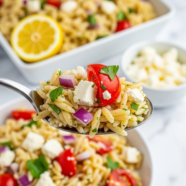 A close-up view of a spoon holding a colorful orzo pasta salad. The salad on the spoon has small, yellow orzo pasta pieces mixed with bright red tomato chunks, small white cheese bits, finely chopped red onion, and green herbs scattered throughout. The background shows more of the salad with similar ingredients, accented with a slice of lemon on the side, all placed on a white marbled surface. The image is sharp with a soft focus on the background, highlighting the textures and colors of the salad. Photo taken with an iphone --ar 4:5 --v 7