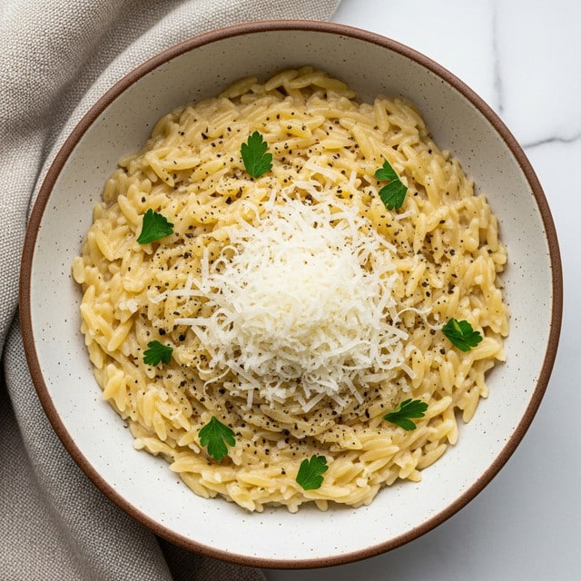A bowl filled with creamy, light yellow orzo pasta, covered with finely grated white cheese in the middle, sprinkled with small black pepper flakes and a few green parsley leaves scattered on top. The bowl is white with a speckled brown edge, sitting on a textured beige cloth against a white marbled surface. photo taken with an iphone --ar 4:5 --v 7