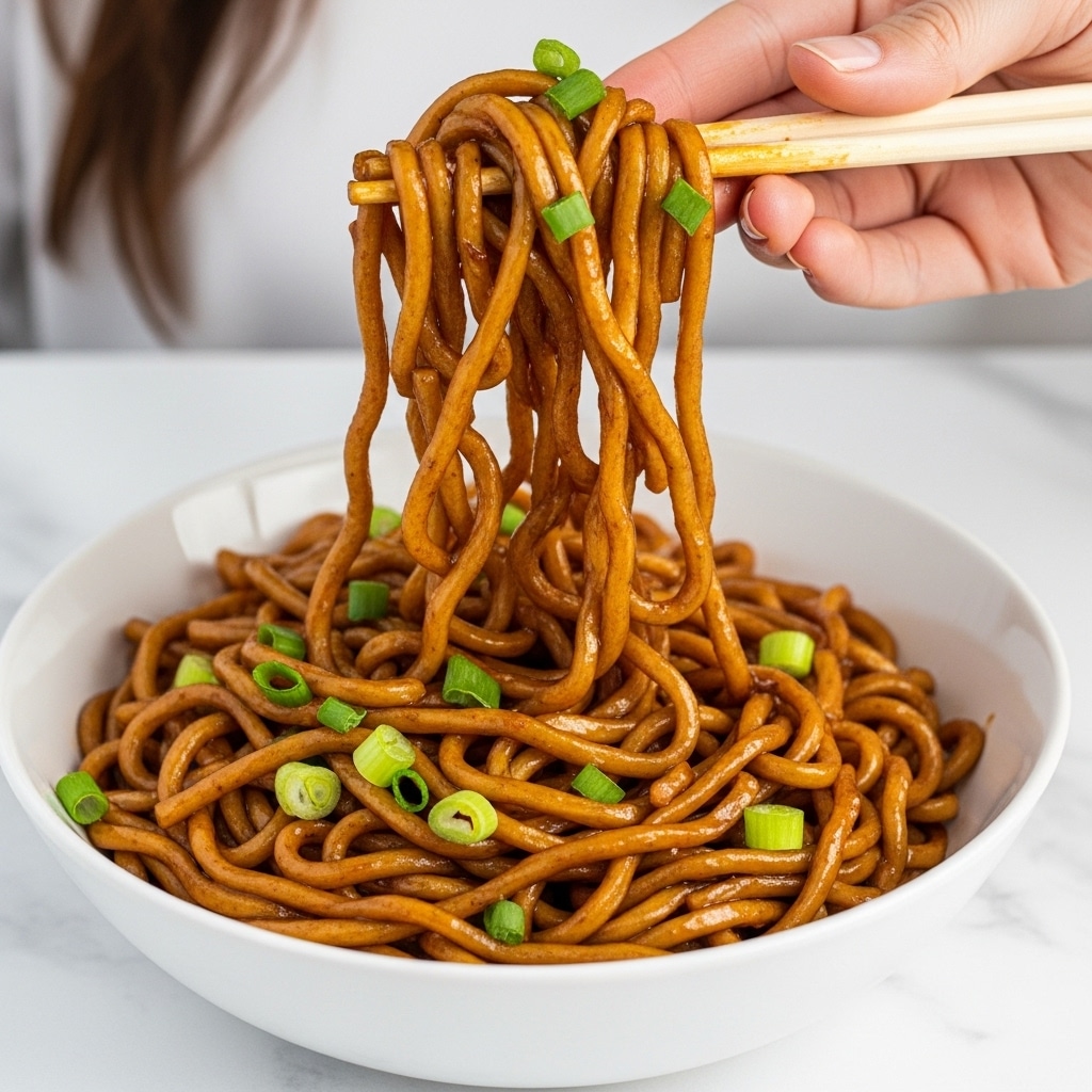 A white bowl filled with a large serving of stir-fried noodles, dark brown in color, glossy with sauce, mixed with small pieces of cooked chicken and green chopped spring onions scattered on top. A woman's hand is lifting a twisted bunch of noodles from the bowl using chopsticks, showing the thick, saucy texture and the rich, shiny coating on the noodles and chicken. The background is a white marbled texture. Photo taken with an iphone --ar 4:5 --v 7