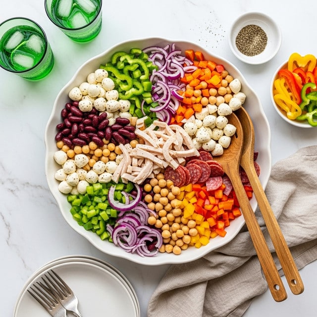 A large white bowl filled with a colorful salad showing multiple layers: dark red kidney beans and light beige chickpeas form a base mixed with small white mozzarella balls, thin slices of reddish-pink salami, and shredded white turkey. Scattered throughout are bright green bell pepper pieces, thin slices of purple onion, small red cherry tomato halves, and bits of orange and yellow bell pepper. Two wooden salad spoons rest inside the bowl on the right side. The bowl is set on a white marbled surface with a green-checked cloth underneath, surrounded by a beige cloth, an empty white plate with silverware, two glasses with ice and green-tinted water, and small white dishes containing sliced bell peppers and ground black pepper. photo taken with an iphone --ar 4:5 --v 7