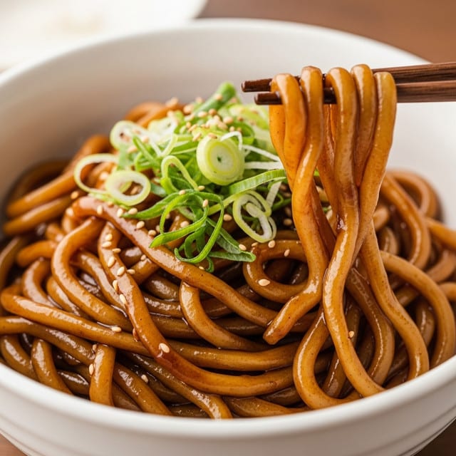 A close-up of a white bowl filled with glossy, dark brown noodles that are thick and coated in sauce. The noodles are mixed with small green sliced onions, adding a fresh pop of color. A woman's hand is holding some noodles lifted above the bowl, showing their texture and the sauce glistening on them. The background is a white marbled surface. Photo taken with an iphone --ar 4:5 --v 7
