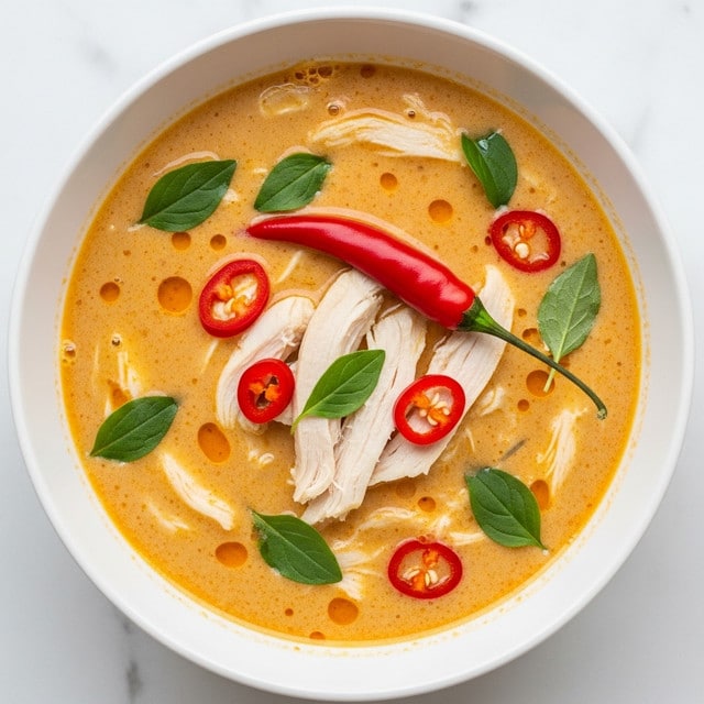A white bowl filled with creamy light orange soup with a smooth texture, featuring several torn pieces of white chicken scattered throughout. Bright red chili peppers sit atop the soup, adding vibrant splashes of color. Small, dark green fresh herb leaves are spread evenly across the surface, providing contrast to the warm soup base. The bowl rests on a white marbled surface, giving a clean and fresh look. photo taken with an iphone --ar 4:5 --v 7