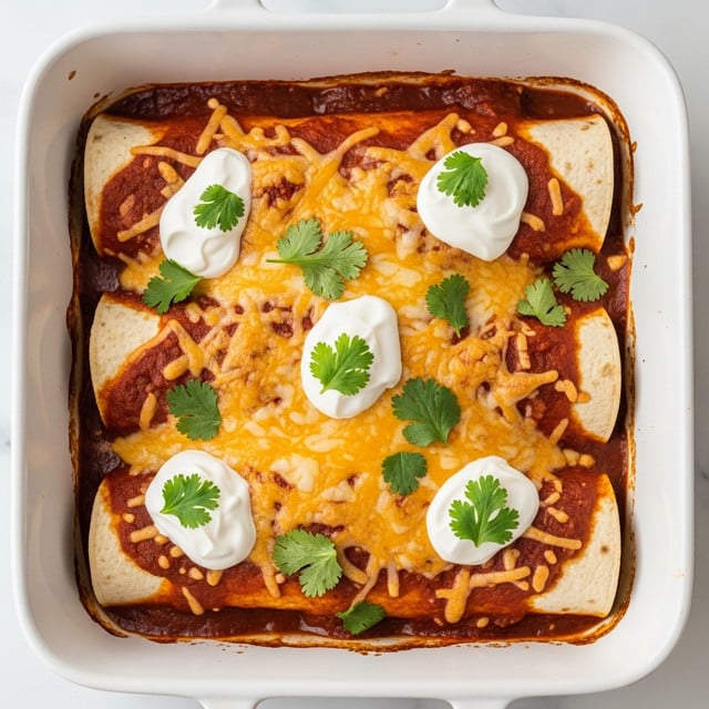 The image shows a white rectangular baking dish filled with two rolled enchiladas placed side by side, topped with melted golden and slightly browned cheese covering the entire surface. On top of the cheese, there is a layer of fresh green cilantro leaves scattered evenly, and dollops of white sour cream spread in spots along the length. The edges of the enchiladas reveal soft tortilla rolls filled with a reddish sauce and a bit of melted cheese inside. The dish rests on a white marbled surface with a gray cloth napkin partially visible on the side. photo taken with an iphone --ar 4:5 --v 7