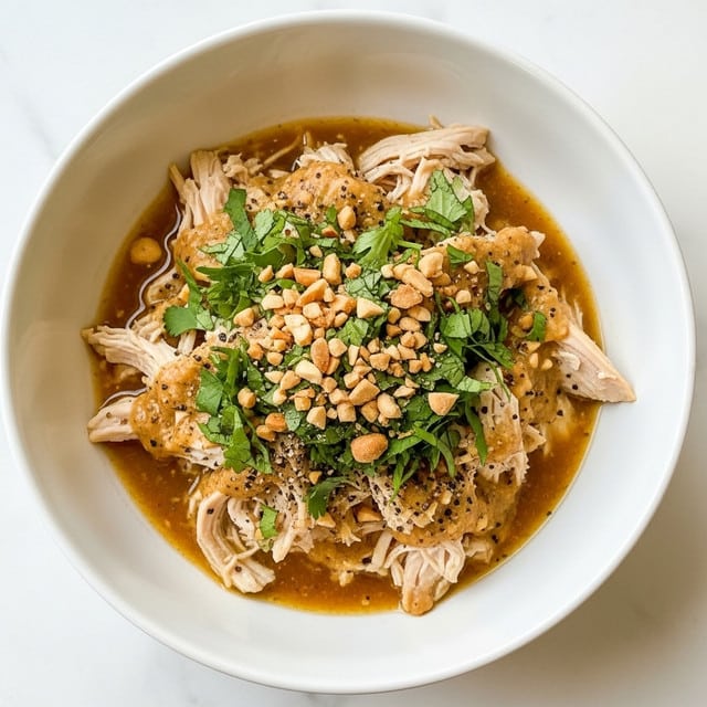 A white bowl filled with shredded chicken pieces covered in a light brown sauce, topped with chopped green herbs and crushed peanuts, creating a mix of green, tan, and beige colors. The chicken looks tender and is sitting in a thin layer of sauce with specks of black pepper and bits of herbs. The bowl rests on a white marbled surface, adding a clean and bright background. photo taken with an iphone --ar 4:5 --v 7