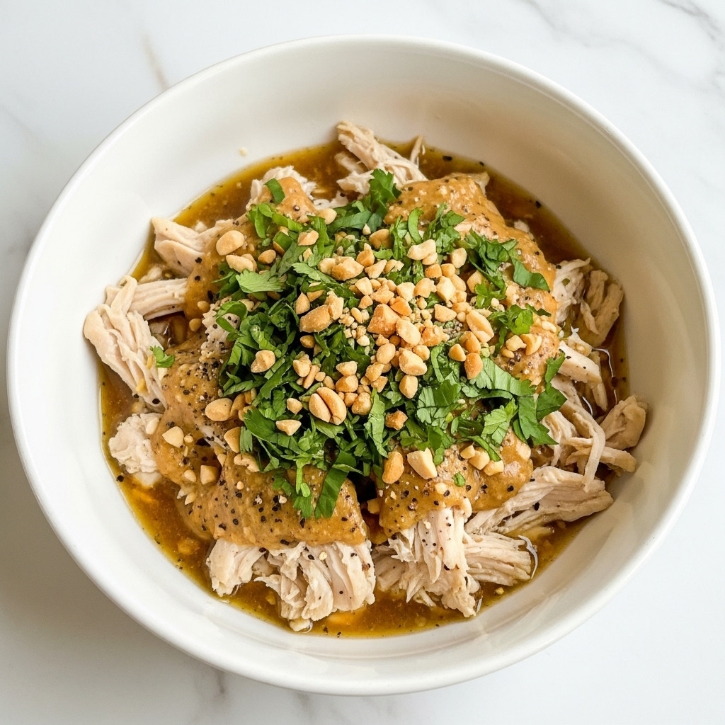 The image shows a white bowl filled with shredded chicken covered in a glossy brown sauce. On top, there are small chopped green herbs and crushed nuts scattered evenly, adding texture and color contrast. The chicken looks tender and juicy with strands clearly separated, while the sauce pools at the bottom, partially soaking the meat. The bowl sits on a white marbled surface, with some green herbs blurred in the background. photo taken with an iphone --ar 4:5 --v 7