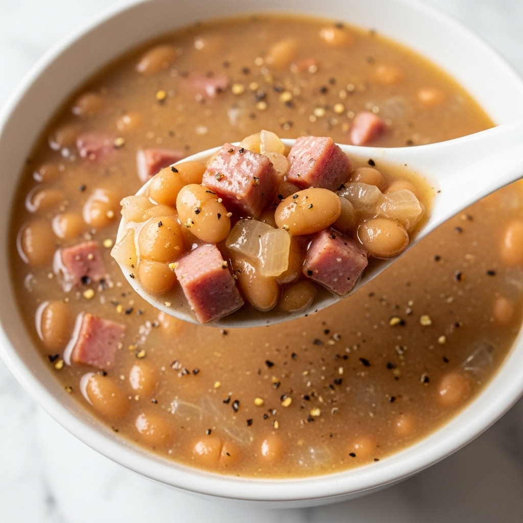 A close-up view of a white spoon lifting a spoonful of warm baked beans mixed with small cubes of ham and translucent cooked onions from a bowl filled with creamy, light brown bean stew. The beans are soft and pale, scattered evenly throughout the thick broth, which has a smooth yet slightly chunky texture. Small black pepper flakes sprinkle lightly across the surface, adding contrast to the soft colors. The bowl rests on a white marbled surface, enhancing the cozy and hearty feeling of the dish. Photo taken with an iphone --ar 4:5 --v 7