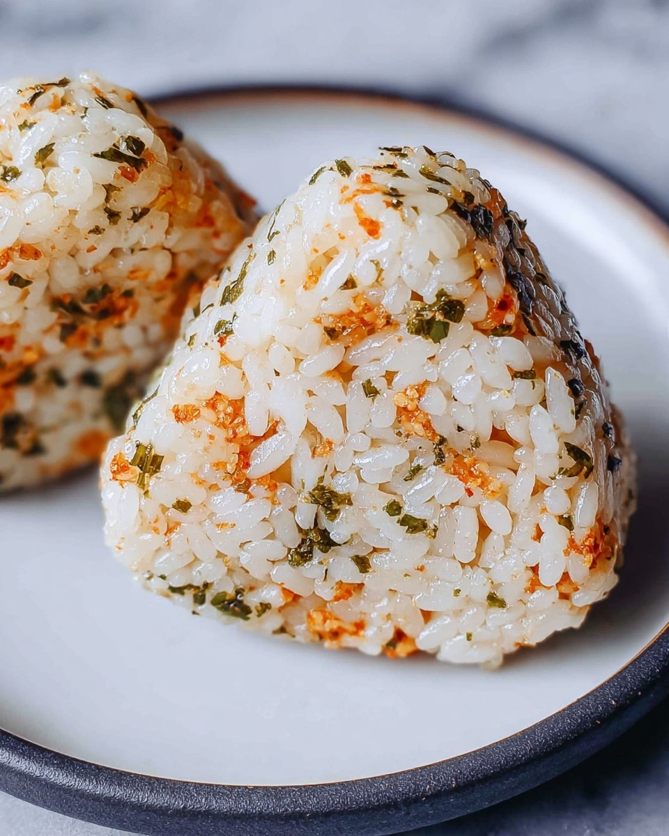 The image shows a close-up of two triangular rice balls on a white plate with a dark rim, placed on a white marbled surface. Each rice ball has two visible layers: the outer layer is white rice mixed with small orange and green bits, likely seasoning or herbs, giving it a speckled look with a slightly glossy texture. The rice grains are distinct and tightly packed, forming a firm shape. The plate's light color contrasts with the vibrant rice balls, highlighting their detailed texture clearly. photo taken with an iphone --ar 4:5 --v 7