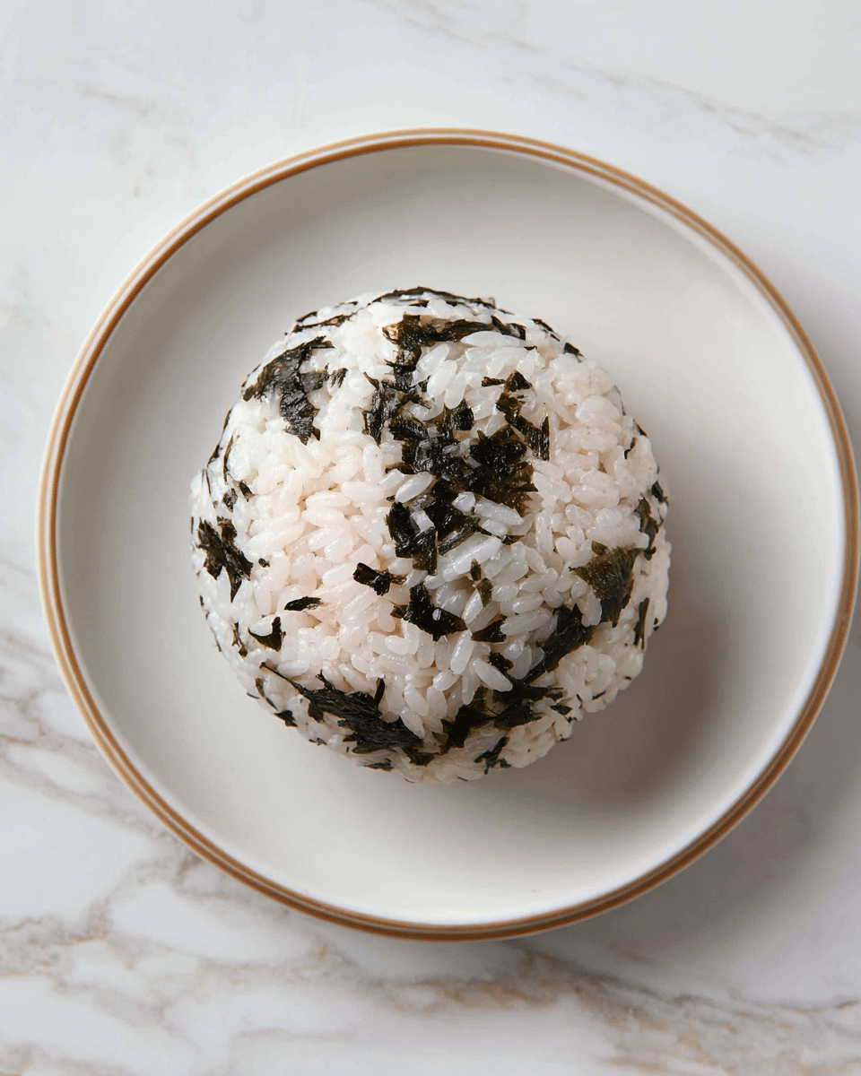 A round rice ball with white rice grains mixed with dark green seaweed flakes on the outside layer, showing a textured look. The inner layer is a soft, light pink filling that looks creamy and smooth. The rice ball is placed in the center of a white plate with a light brown rim, all set on a white marbled texture surface. Photo taken with an iphone --ar 4:5 --v 7