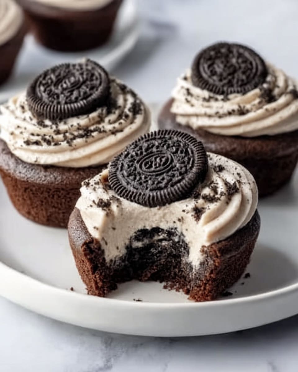 Three chocolate cupcakes sit on a white plate placed on a white marbled surface. Each cupcake has a dark brown chocolate base with a thick layer of cream cheese frosting on top, which is white with small black cookie crumbs mixed in. On the very top center of each cupcake, there is a whole Oreo cookie pressed slightly into the frosting. One cupcake at the front has a bite taken out of it, revealing the moist chocolate cake inside and the creamy white frosting layer just beneath the cookie. The image is well-lit, focusing on the texture and details of the cupcakes, photo taken with an iphone --ar 4:5 --v 7