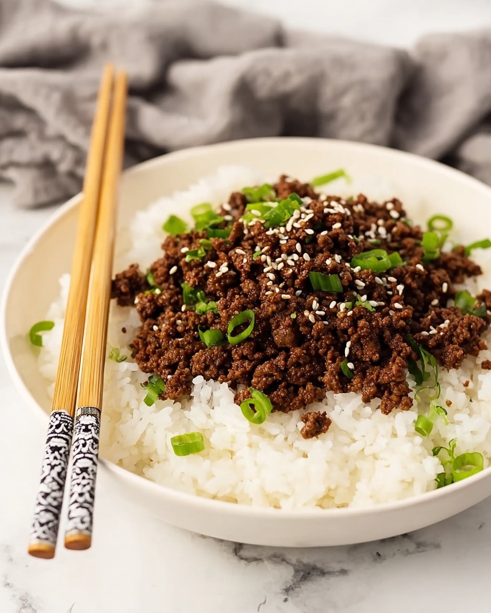 A white shallow bowl contains two main layers: at the bottom, there is a fluffy layer of white rice with a soft and slightly sticky texture, and on top, there is a generous layer of cooked ground beef that is dark brown and crumbly. The beef is garnished with small white sesame seeds and bright green chopped scallions, adding a fresh look. A pair of light wooden chopsticks rests on the edge of the bowl, positioned diagonally. The bowl sits on a white marbled surface with a soft gray cloth in the background. photo taken with an iphone --ar 4:5 --v 7