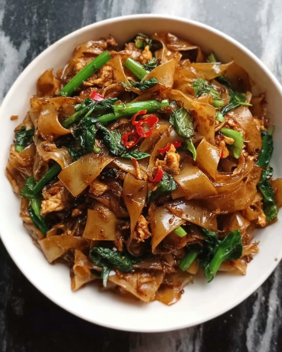This image shows a black bowl filled with stir-fried wide flat noodles mixed with green vegetable pieces and small bits of browned meat. The noodles have a shiny, dark brown color from the sauce, and the green vegetables stand out against the rich color of the noodles. The texture looks soft and slightly oily, with the vegetables adding a fresh, crisp touch. The bowl is placed on a white marbled surface. Photo taken with an iphone --ar 4:5 --v 7