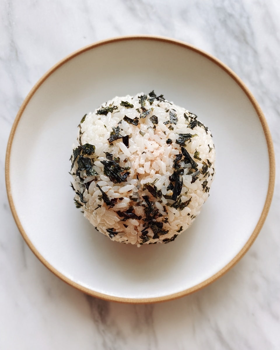 A close-up of a round rice ball with three visible layers: the outer layer is made of sticky white rice mixed with dark green seaweed pieces, wrapping fully around the middle layer which is light beige and has a soft, finely textured filling, likely tuna or a similar mixture. The rice ball is placed on a white plate with a slightly speckled edge and the scene rests on a white marbled surface. photo taken with an iphone --ar 4:5 --v 7