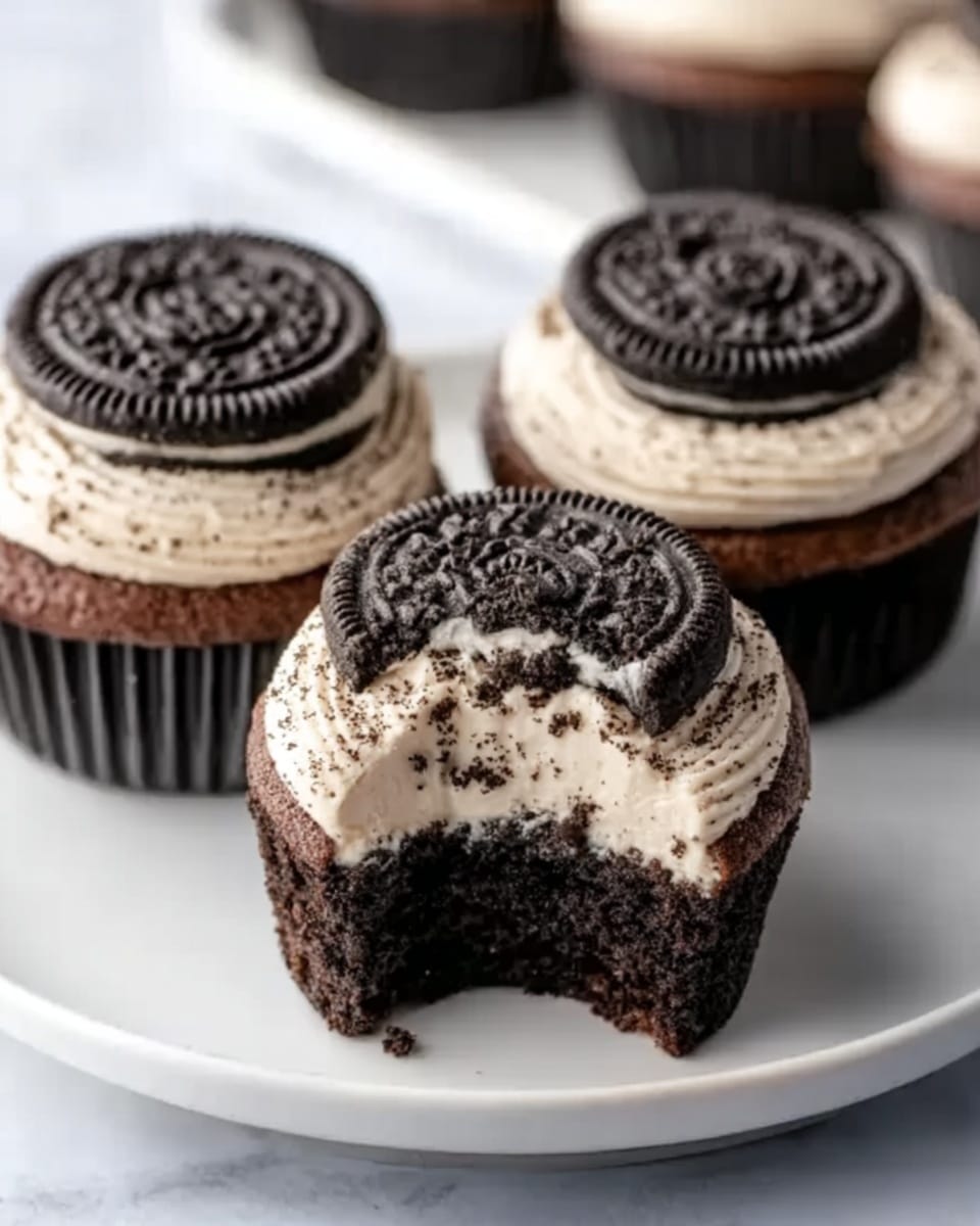 The image shows three chocolate cupcakes on a white plate, each topped with creamy white frosting that has small black cookie crumbs mixed in. On top of each cupcake’s frosting layer, there is a whole chocolate sandwich cookie with a dark textured surface. One cupcake is cut in half to show a soft, moist chocolate cake inside, with a bitten part revealing the creamy frosting and cookie on top. The plate sits on a white marbled surface, and some cookie crumbs are scattered around the cupcakes. Photo taken with an iphone --ar 4:5 --v 7