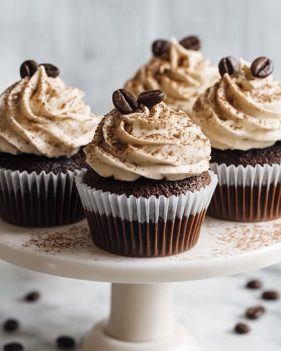 The image shows four dark chocolate cupcakes placed on a white cake stand with a white marbled background. Each cupcake has a rich, dark brown base with a moist texture. On top, there is a swirl of creamy light beige frosting dusted with cocoa powder. Each frosting swirl is topped with a single dark coffee bean as decoration. Some loose coffee beans are scattered on the stand near the cupcakes. A woman's hand with a smooth skin tone is delicately touching one of the cupcakes on the right side. The overall look is neat and inviting, with soft natural light enhancing the textures and colors. photo taken with an iphone --ar 4:5 --v 7