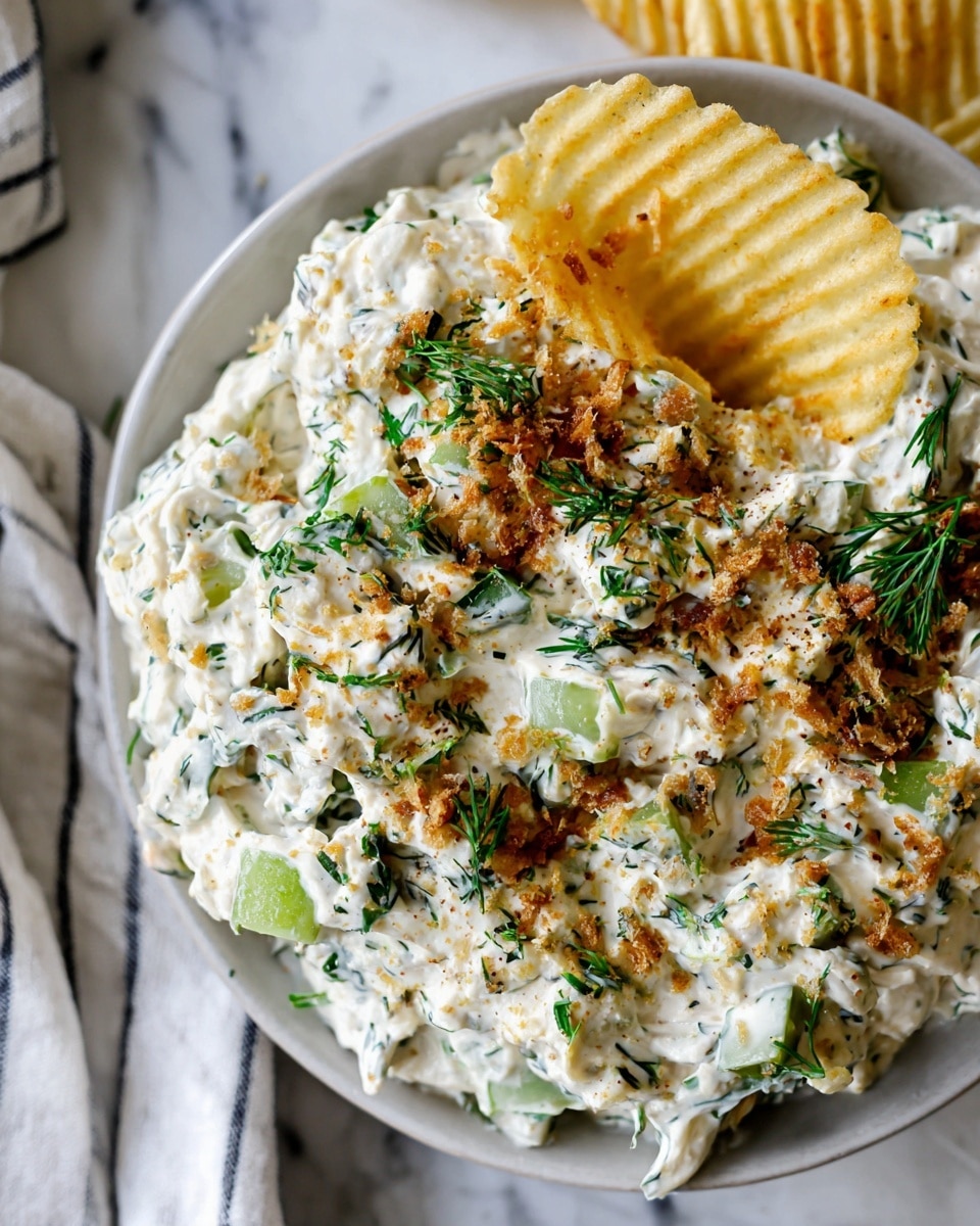 A white round bowl filled with a creamy, chunky dip made of white cheese mixed with small pieces of green and white pickles, chopped green herbs, and sprinkled with fine brown crumbs and fresh green herb leaves on top. A single crinkle-cut yellow chip is partly dipped into the creamy mixture, resting on the bowl’s edge. The bowl sits on a white marbled surface, next to a white cloth with dark stripes, with a few more yellow chips visible around the bowl. photo taken with an iphone --ar 4:5 --v 7