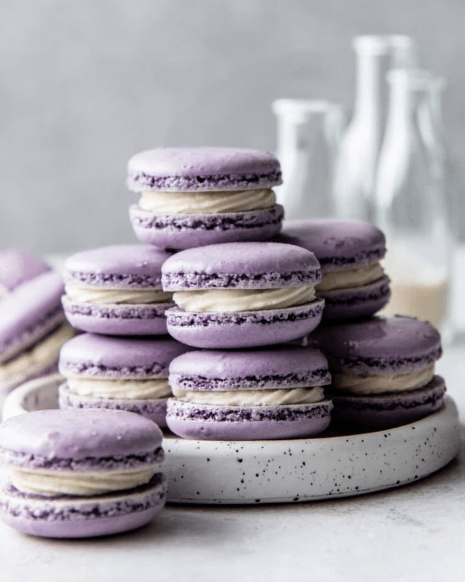 A stack of soft purple macarons with thick pale cream filling sits on a round white plate with small black dots. The macarons have smooth, slightly shiny tops and a textured middle layer from the filling. Two macarons lie next to the plate on a white marbled surface. In the blurry background, there are two clear glass bottles, one taller and one shorter. The image looks neat and bright, showing the delicate details of the macarons. Photo taken with an iphone --ar 4:5 --v 7