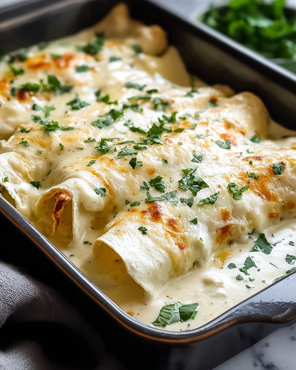 A close-up view of a rectangular dark baking dish filled with three rolled crepes covered in a creamy white sauce with a slightly browned, melted cheese layer on top. The crepes show soft, folded edges and are evenly coated with the sauce, sprinkled with fresh green chopped herbs. The creamy sauce looks smooth and thick, pooling gently around the crepes inside the dish. The dish is set on a white marbled textured surface with a soft focus in the background. photo taken with an iphone --ar 4:5 --v 7