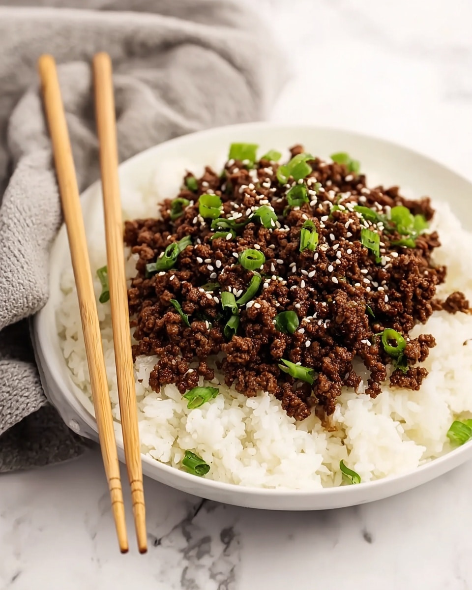 A white bowl filled with a base layer of fluffy white rice, topped with a generous layer of cooked minced beef that is dark brown and textured with small chunks. The beef is sprinkled with white sesame seeds and small chopped green onions, adding contrast with their bright green color. Two light brown wooden chopsticks rest on the edge of the bowl. The dish is set on a white marbled surface, with a soft grey cloth casually placed in the background. photo taken with an iphone --ar 4:5 --v 7